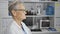 Serious-faced grey-haired senior woman scientist, absorbed in a lab experiment, looking to the side in a busy laboratory