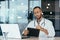 Serious and focused hispanic doctor in medical coat and stethoscope behind paperwork, man in glasses working inside