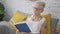 Serious grey-haired senior woman comfortably sitting on her room\\\'s bed, doubting and thinking as she reads a book in her cosy