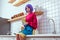 serious housewife with purple hair and colorful clothes sitting on kitchen counter and holding glass jar