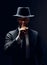 Serious man in black suit and hat keeps finger on lips, making hush gesture and keep conspiracy isolated on dark background