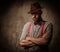 Serious old-fashioned man with hat wearing suspenders and bow tie, posing on dark background.