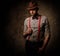 Serious old-fashioned man with hat wearing suspenders and bow tie, posing on dark background.