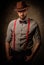 Serious old-fashioned man with hat wearing suspenders and bow tie, posing on dark background.