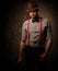 Serious old-fashioned man with hat wearing suspenders and bow tie, posing on dark background.