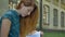 Serious pensive young ginger woman reading book and looking at camera, sitting on street with building in background