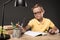 serious schoolboy in eyeglasses doing homework at table with colour pencils books lamp and textbook on grey