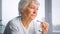 Serious senior businesswoman with short grey hair holds glasses in trembling hand against blurred window at home