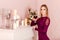 A serious slender twenty-year-old woman stands by the fireplace and holds a glass of red wine in her hand