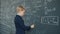 Serious small kid studying in university indoors writing formulas on chalkboard