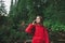Serious tourist girl speaking on the phone looking far ahead in the mountain fir forest. Female hiker in red raincoat is making a