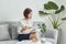 Serious young Asian girl in blouse and skirt.sitting on sofa and drinking tea while reading book in living room