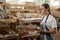 Serious young baker with tablet standing by display with fresh baked bread, smiling woman in supermarket bakery