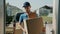 Serious young deliveryman bringing boxes from van to house working in suburban area