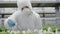 Serious young female scientist in respirator and protective workrobe adding pesticides to plants in greenhouse. Portrait