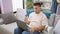 Serious young hispanic guy engrossed in online payment, comfortably resting on living room sofa, laptop poised