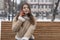 Serious young woman with gadget. Brunette girl in sheepskin coat sits on bench in winter park and talks on smartphone