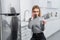 Serious young woman holding card with lettering home inspection and talking on smartphone near broken refrigerator