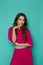 Serious young woman in magenta pleated dress is posing with hand on chin