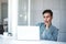 Seriously Young Businessman Working on Computer Laptop in Office. Hand on Shin, Sitting on Desk with Thoughtful Posture.