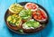 A set of crostini with feta, ricotta, vegetables and fruits and fresh herbs on a blue background.