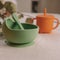 Set of silicone baby dishes. Green bowl, spoon and orange sippy cup on table against eucalyptus sprig. Square frame