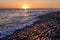 Setting sun on the horizon is reflected in the water and a pebble beach