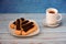 Several chocolate-coated cream eclairs on a plate and a cup of hot cappuccino on a wooden table