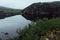 Severe north grey arctic landscape slope of granite rocks and quiet dark lake with lush green meadow in haze in overcast weather.