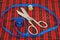 Sewing tools: large scissors and thread with needles lying on a checkered red cloth surrounded by a blue measuring tape, front