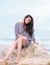 Sexy Caucasian Woman Outdoors, Sitting on a Rock, Near the Sea, in a Marine Beach Dress, with Long Curly Hair and Sad Attitude