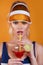 Sexy woman in swimsuit and trendy plastic visor holding glass of fresh drink on yellow background