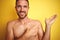 Sexy young shirtless man showing nude fitness chest over yellow isolated background amazed and smiling to the camera while