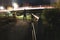 A shallow depth of field, shot of a hooded figure in front of street lights and traffic trails at night in a park