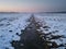 A shallow ditch with frozen water cuts across a meadow under snow at dusk, a mist in the distance and a purple glow in the sky