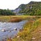 A shallow fast mountain river with a rocky bottom. In the background there are rocks overgrown with forest