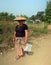 A Shan woman wearing a traditional straw hat walking on unpaved road with a plastic bag in her hand