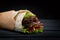 Shawarma rolled in lavash, moist grilled meat with onion, herbs and vegetables on wooden black background.