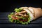Shawarma rolled in lavash, moist grilled meat with onion, herbs and vegetables on wooden black background.