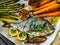 Sheet pan dinner - roasted whole sea bream with asparagus, carrots, lemon ,rosemary and garlic on cooking pan on wooden table