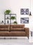Shelf with green plants behind big comfortable leather sofa in white living room with industrial posters