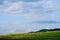 A shepherd grazes a flock of sheep on a green hillside, against a blue sky with clouds.