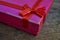 Shiny closed pink gift box with red ribbon and golden stitching on the wooden background