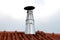 Shiny metal small chimney with leaning protective cover on top of family house roof surrounded with roof tiles