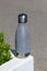 Shiny metal thermos drinking bottle with partially rough texture left on edge of white concrete flower pot on public sidewalk