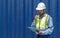 Shipment worker with safety vest and hardhat looking at clipboard while holding walkie talkie. A large steel cargo container is