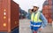 Shipment worker with safety vest and hardhat standing with mobile phone in his hand. A large steel cargo containers stacked in the