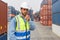 Shipment worker with safety vest and hardhat standing with walkie talkie in his hand. A large steel cargo containers stacked in