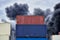 Shipping containers stacked in storage with plumes of black toxic smoke from a fire against a blue sky.