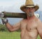 A Shirtless Cowboy Shoulders a Fence Post Driver